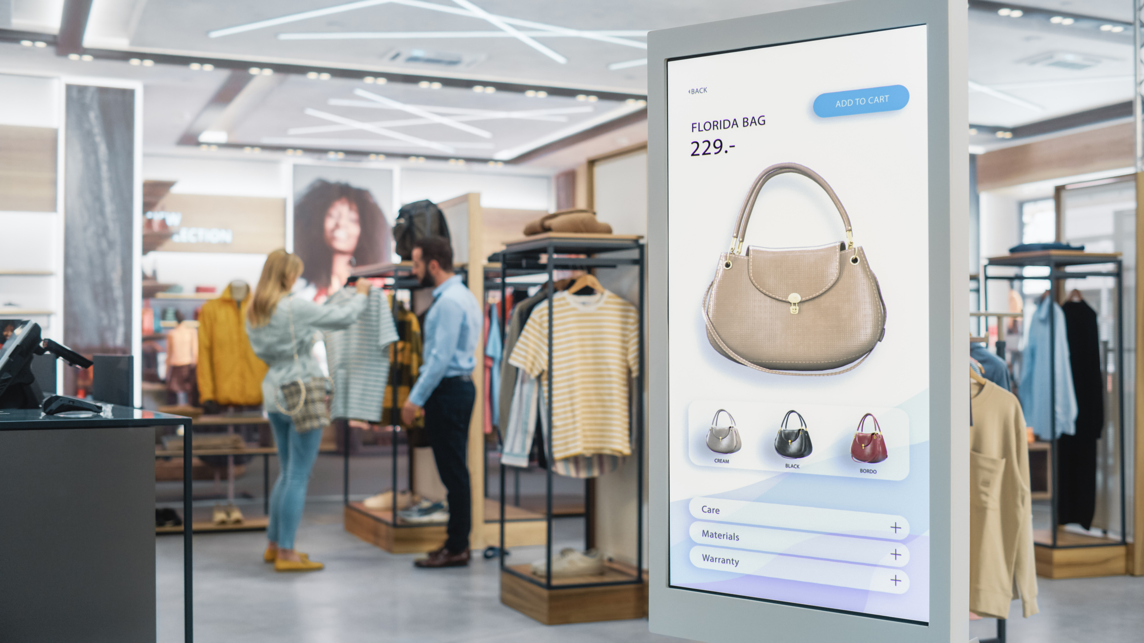 Shot of a Floor-Standing LCD Touch Screen Display with User Interface of Online Clothing Shop Standing in Clothing Store Self service Checkout with Hand Bag Diverse People in Shop Buying Clothes - WPG Americas Inc.